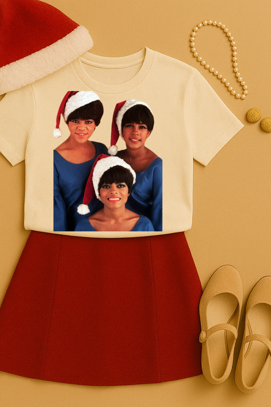 
Cream colored unisex t-shirt featuring The Supremes in blue dresses wearing red and white Santa hats in a festive holiday design. Shirt is styled with red midi skirt, cream Mary Jane shoes, pearl necklace, gold earrings, and Santa hat on a warm beige background.